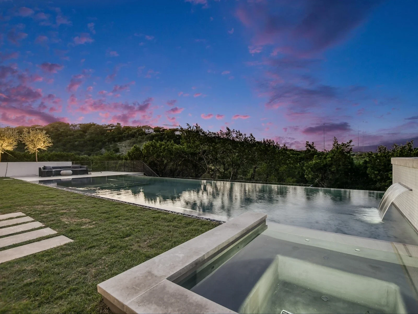 Infinity-edge pool reflecting a pink twilight sky over wooded hills beside a modern stone home