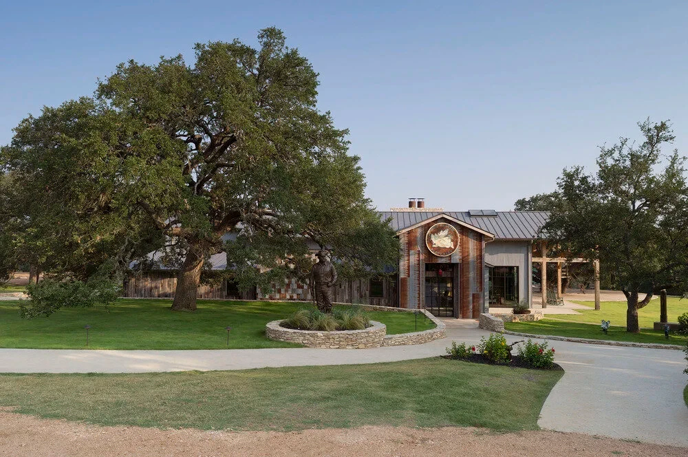 Timber-and-stone lodge with a metal roof set among live oaks along a curving driveway at dusk