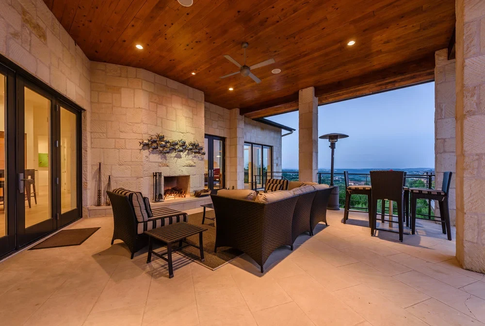 Covered stone patio with a wood ceiling, fireplace, and wicker seating overlooking the Hill Country at dusk