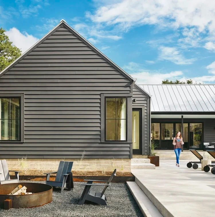 Gray clapboard home with a metal roof, Adirondack chairs around a steel fire pit on a gravel patio