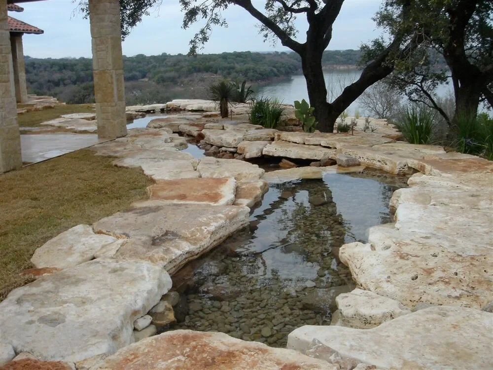 Stone-edged stream running past limestone slabs and oaks toward a lake in the Hill Country