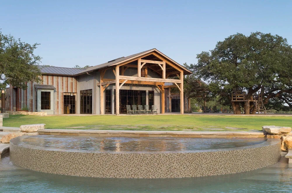 Timber-framed lodge with covered porch behind a still pebble-edged pool, framed by oaks