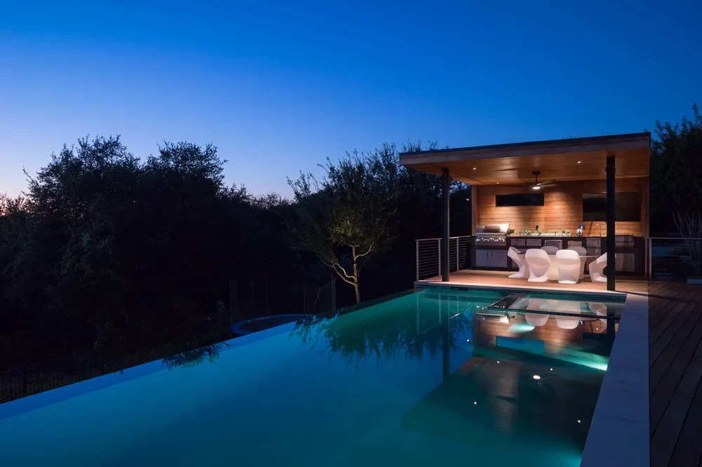 Geometric infinity pool glowing at dusk beside a wood pavilion outdoor kitchen above dark Hill Country trees